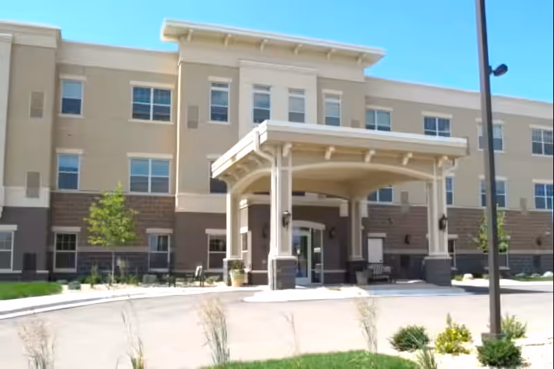 Front exterior view of a three-story senior living facility building with a covered entrance, windows, small trees, and landscaping under a clear blue sky.