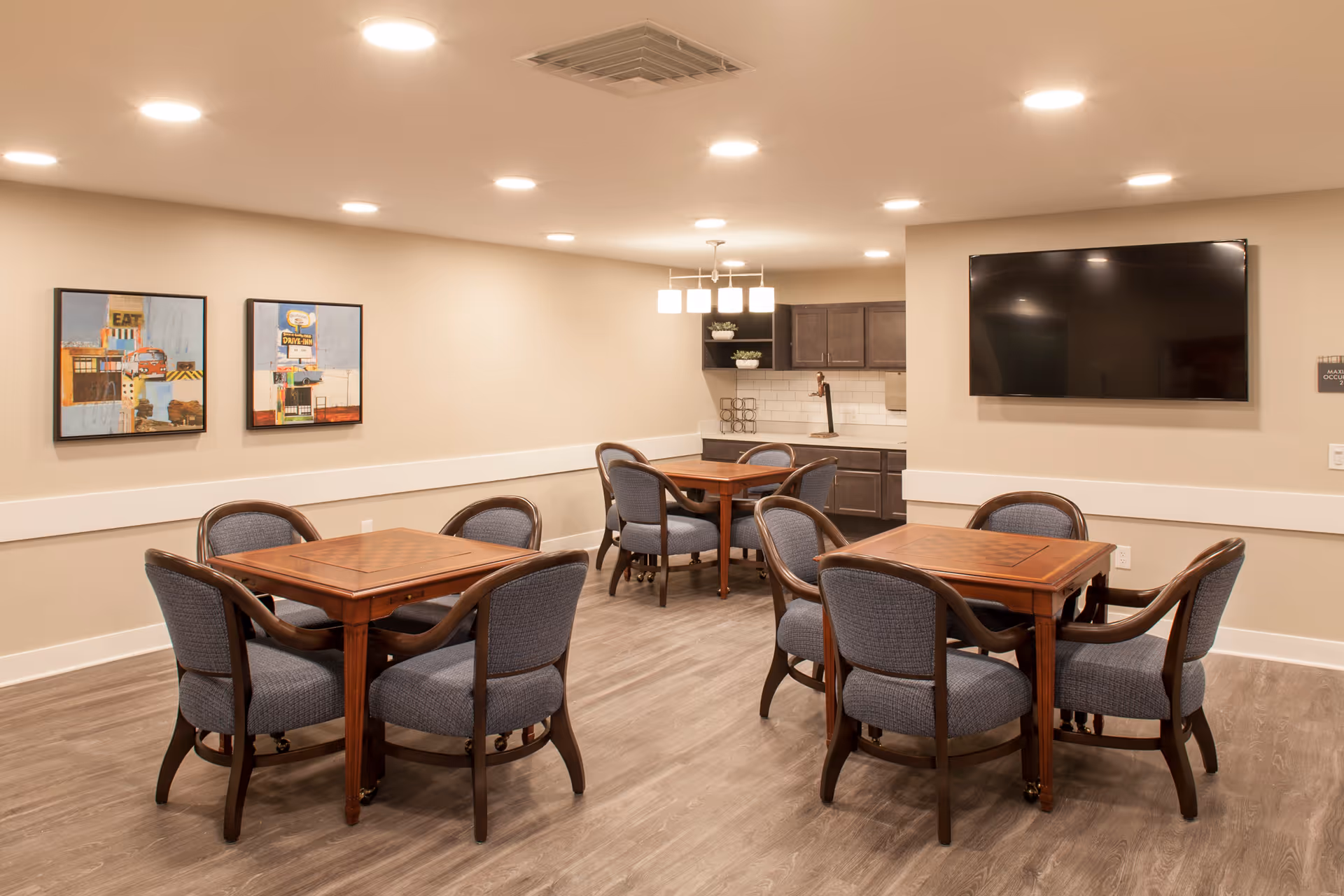 A bright communal dining/activity room with wooden tables and upholstered chairs, a small kitchenette and a wall-mounted TV.