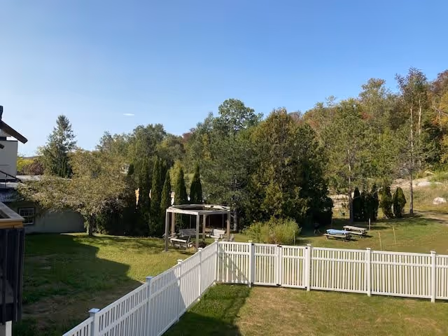 Outdoor view of a senior living facility's fenced yard with green grass, trees, and a wooden pergola with benches underneath. There are picnic tables and a backdrop of more trees and a clear blue sky.