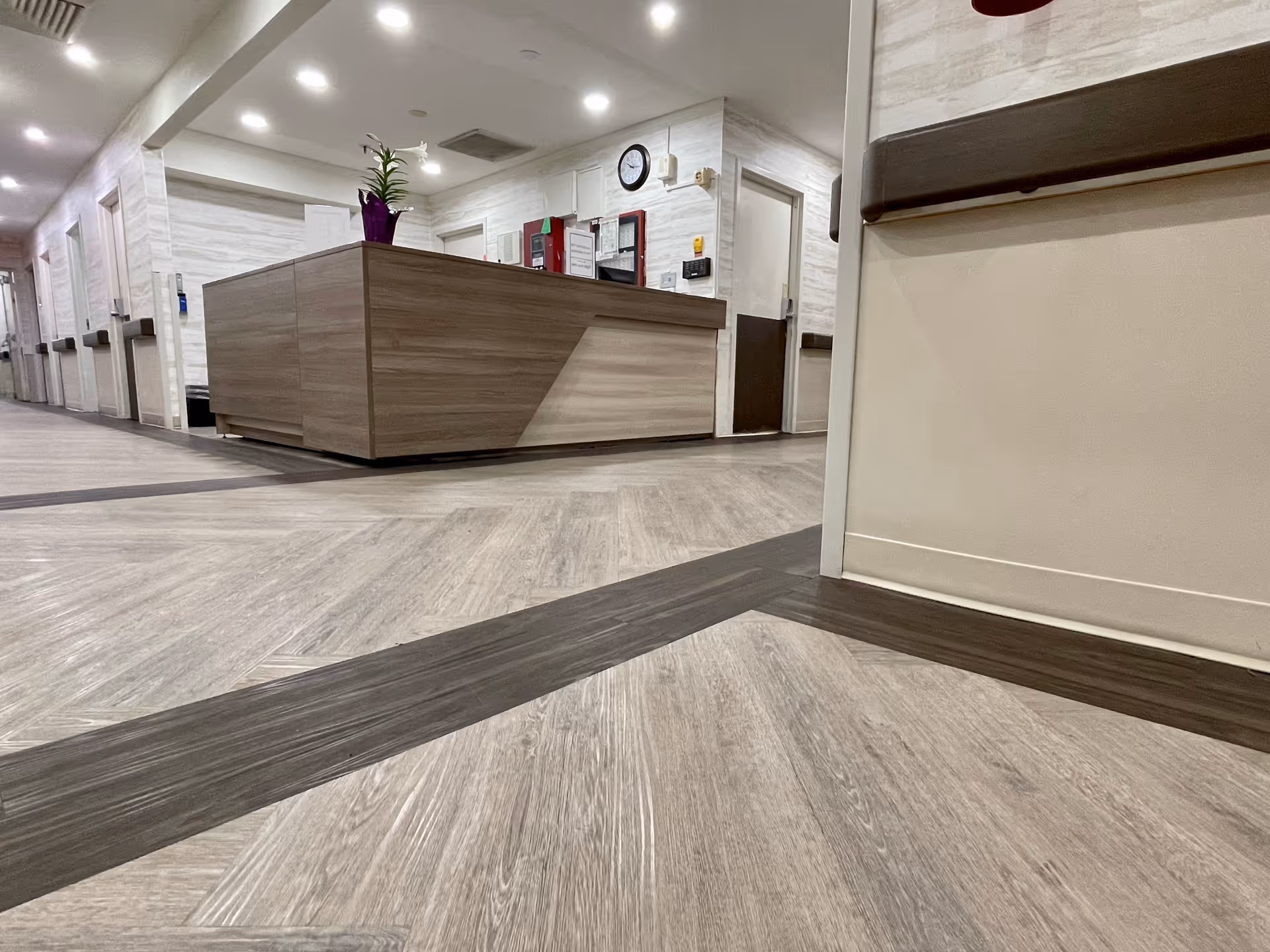 Low-angle view of a reception desk and corridor inside a senior living facility with wood-look flooring and wall handrails.