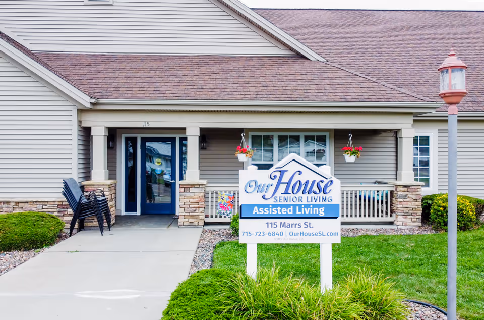 Front entrance of Our House Senior Living assisted living facility with a sign displaying the name, address, phone number, and website. The building has beige siding, a brown shingled roof, a blue door, and a small porch with hanging flower pots and stacked chairs.