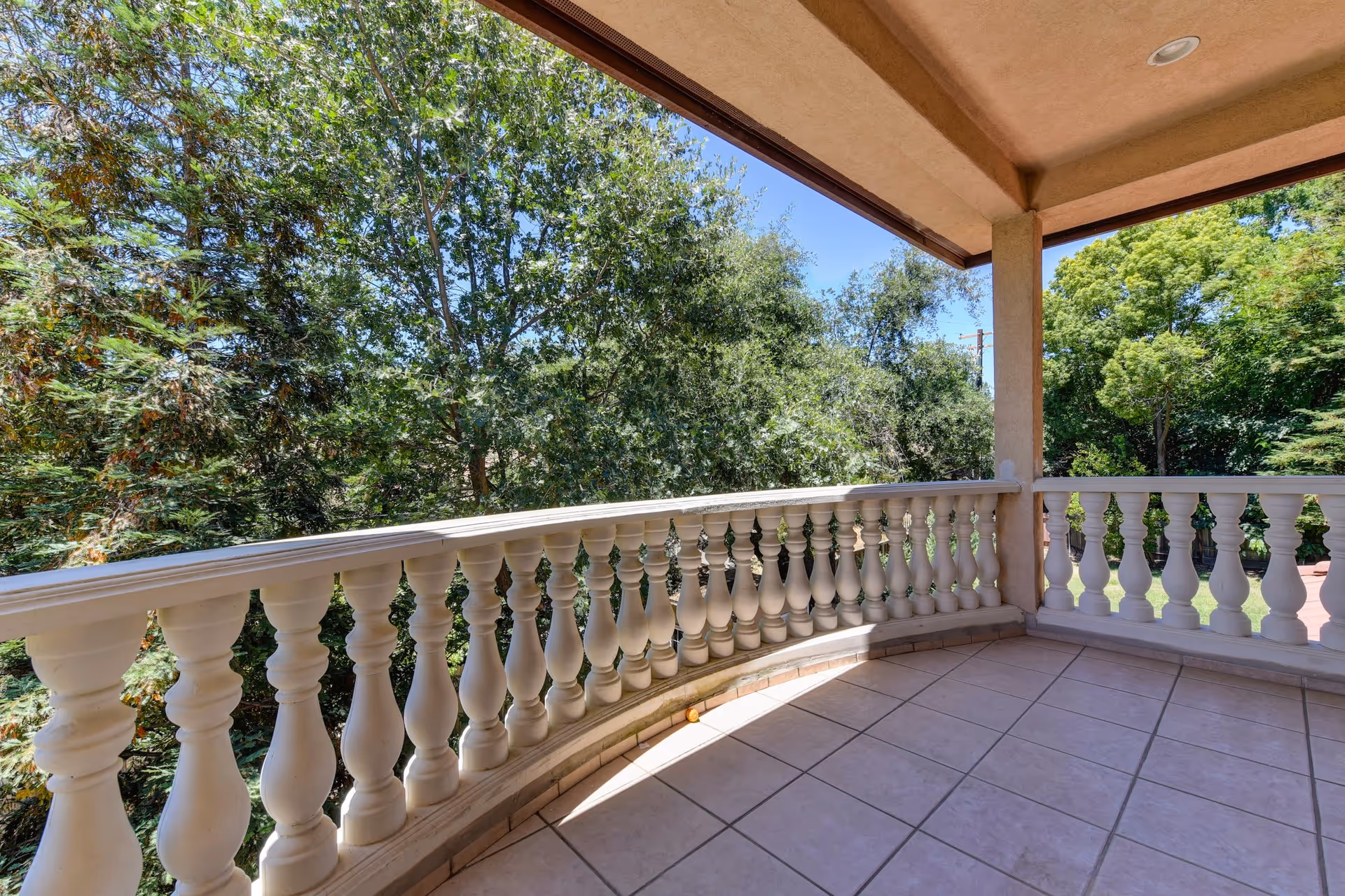A covered outdoor balcony with white balustrade railing overlooking lush green trees under a clear blue sky. The balcony floor is tiled and the ceiling has recessed lighting.