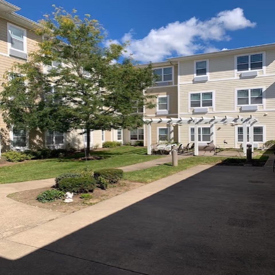 Outdoor courtyard area of a senior living facility with a beige three-story building surrounding a green lawn, a tree, bushes, a white pergola with chairs underneath, and a clear blue sky with some clouds.