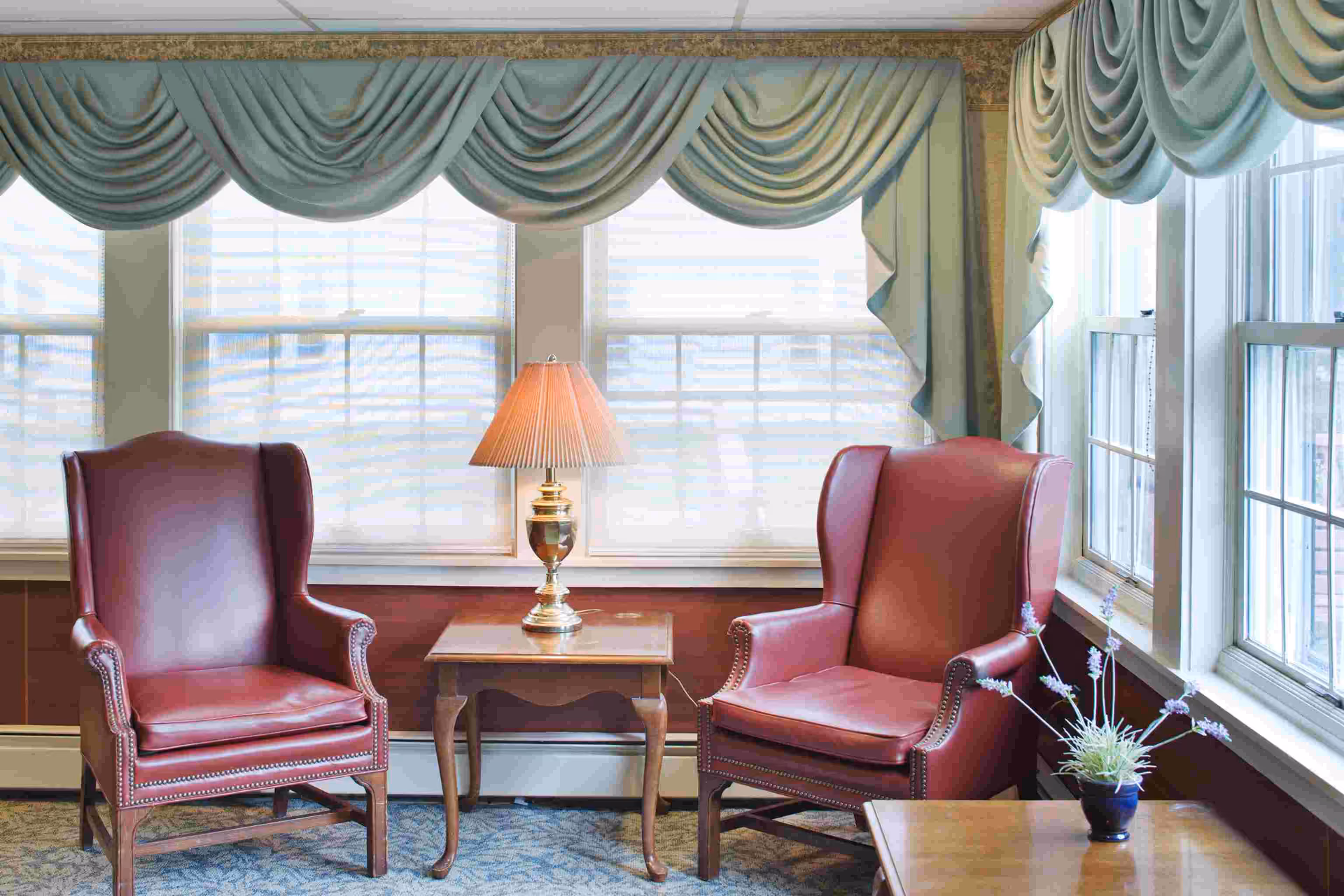 Two red leather wingback chairs flank a wooden side table with a lamp in a sunlit windowed sitting area.