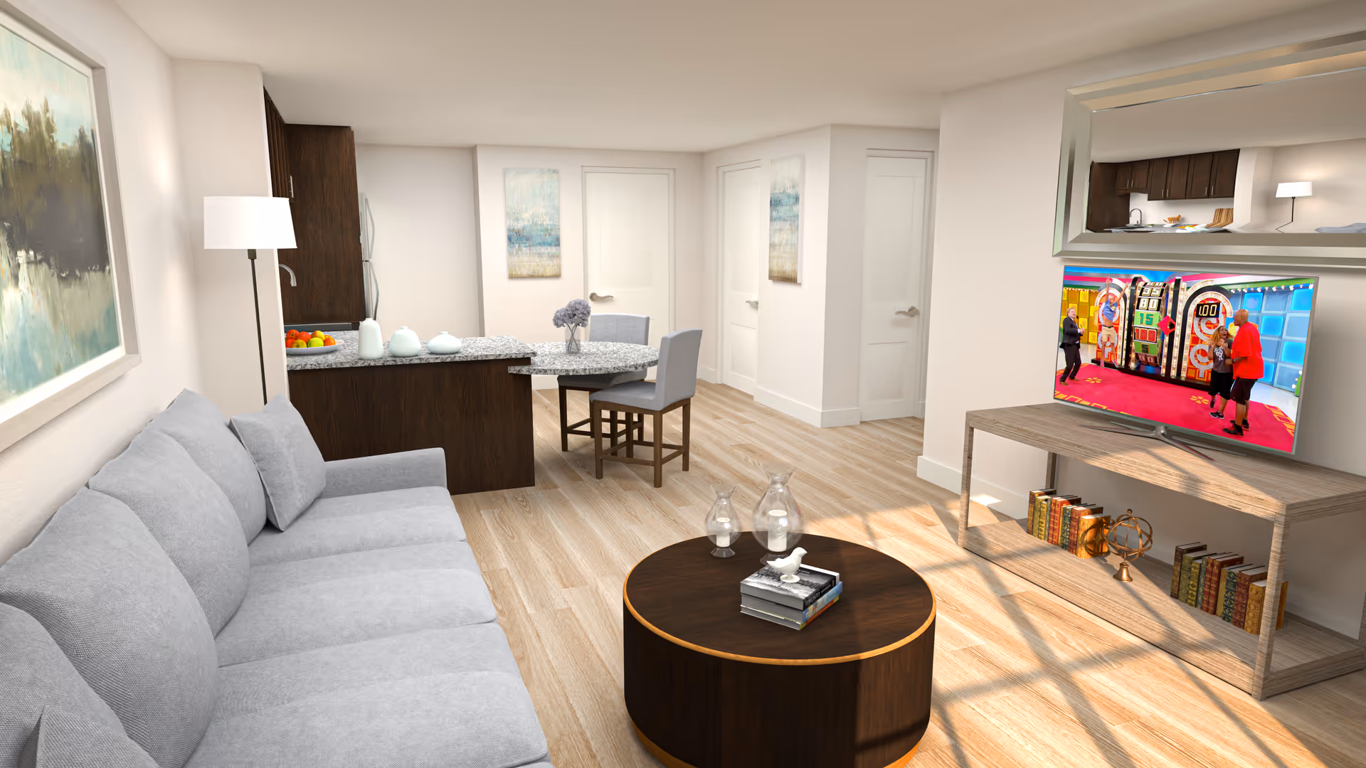 A modern living room with a light gray sofa, a round dark wood coffee table with decorative items, a flat-screen TV on a wooden stand showing a game show, a small dining area with a round table and two chairs, and a kitchen area with dark wood cabinets and granite countertops. The room has light wood flooring and white walls with framed artwork.