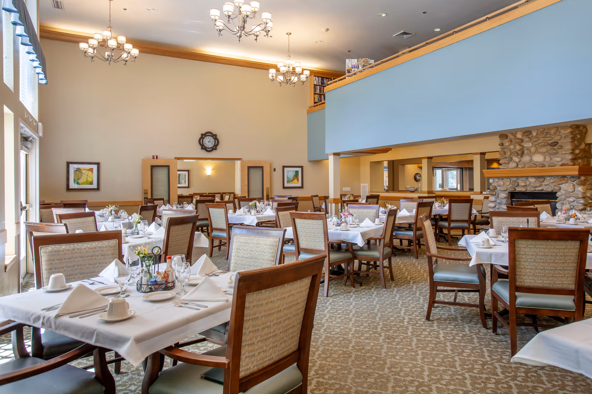 A spacious dining room with multiple tables covered in white tablecloths, set with plates, cups, napkins, and silverware. The room features wooden chairs with patterned upholstery, a stone fireplace on the right, chandeliers hanging from the ceiling, and large windows on the left letting in natural light. There is a blue half-wall balcony area above and framed artwork on the walls.