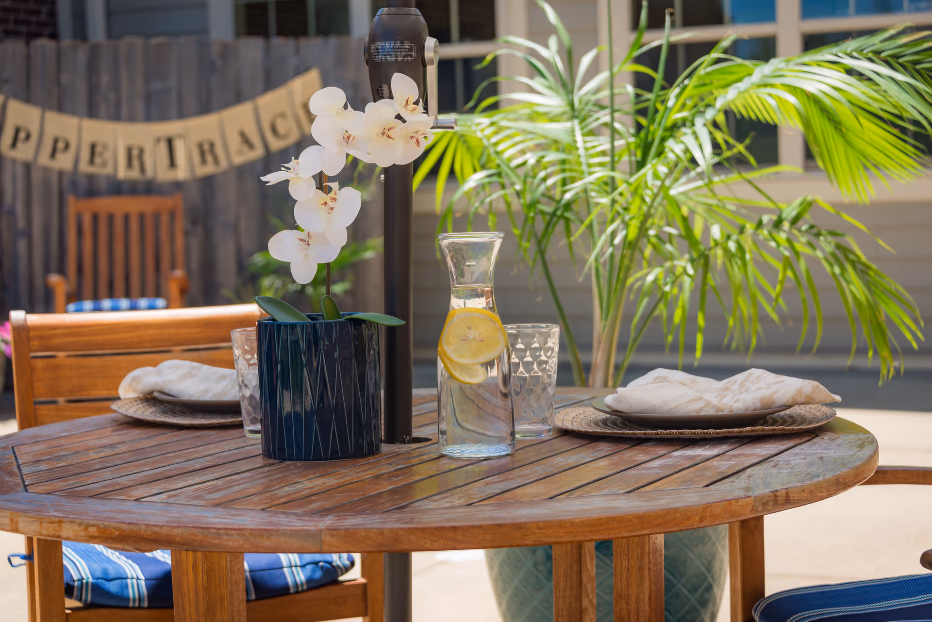 Outdoor wooden patio table set with plates, a blue vase of white orchids, and a carafe of lemon water, with potted plants and a 'Copper Trace' banner in the background.