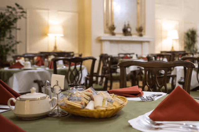 A dining room with tables set for a meal, featuring green tablecloths, red folded napkins, a basket with condiments, a teapot, and silverware. The room has wooden chairs, warm lighting from lamps, and a fireplace in the background.