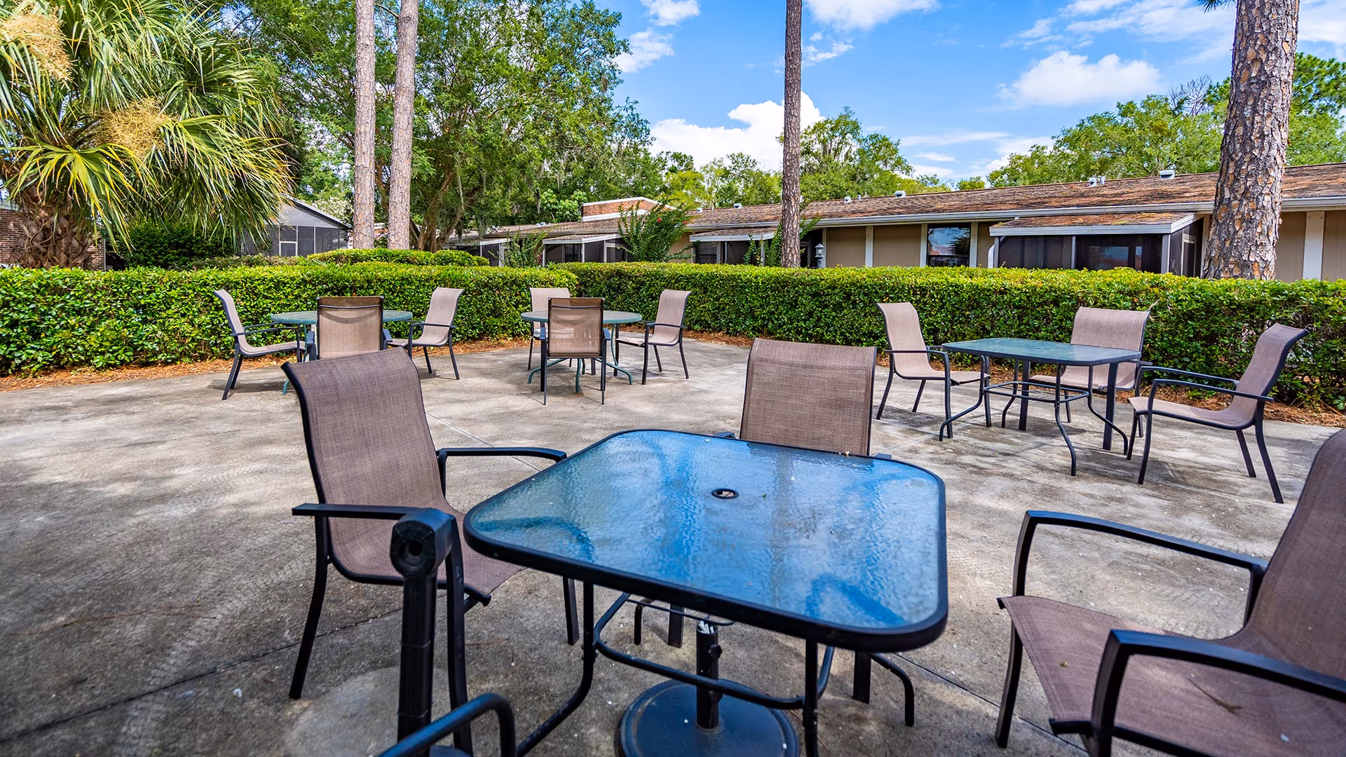 Outdoor patio area with several glass-top tables and brown mesh chairs arranged on a concrete surface. The patio is surrounded by green hedges and tall trees, with a single-story building visible in the background under a partly cloudy blue sky.