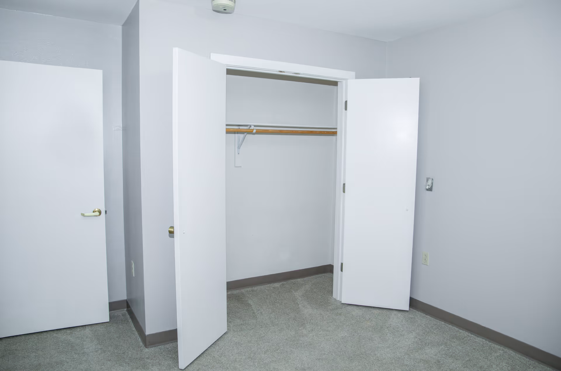 Empty room with light gray walls and carpeted floor, featuring an open closet with white double doors and a wooden hanging rod inside. There is also a closed white door with a brass handle on the left side of the image.