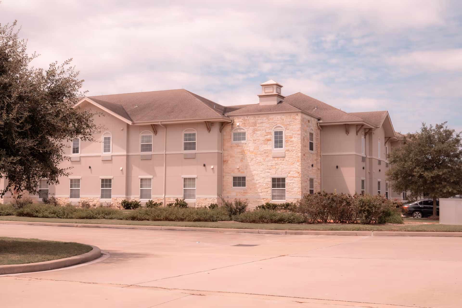 Exterior view of a two-story senior living facility building with beige and stone facade, multiple windows, surrounded by trees and shrubs under a partly cloudy sky.