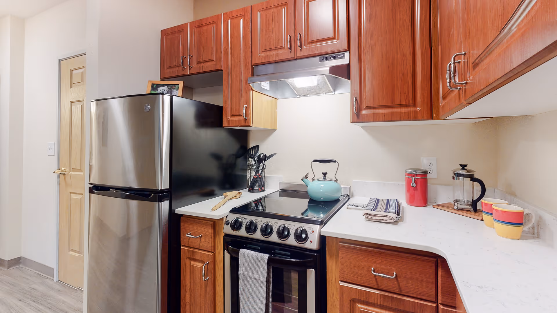 Small kitchen featuring a stainless steel refrigerator, a stove with a teal kettle, wooden cabinets and white countertops.