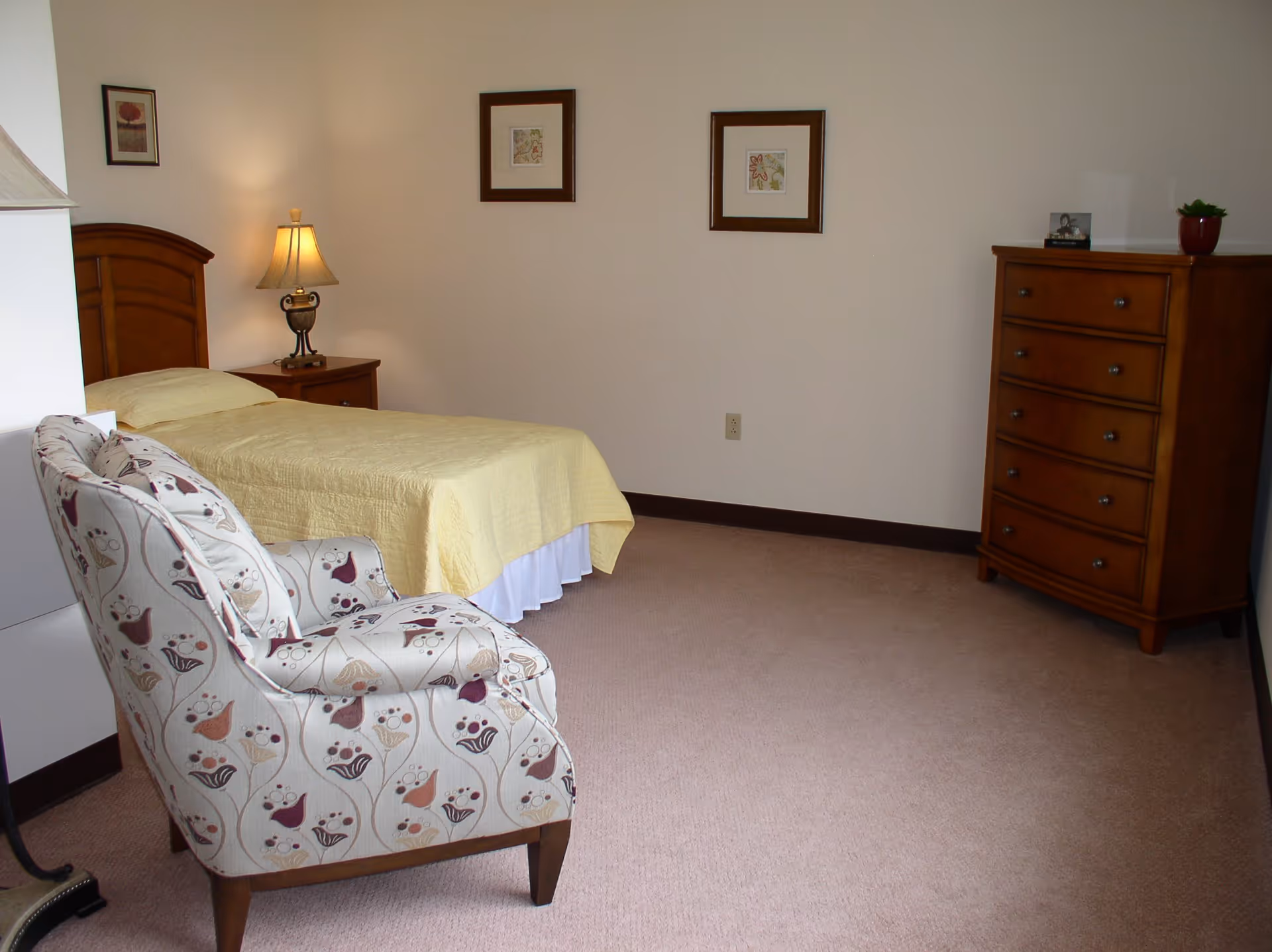 A bedroom with a single bed covered in a yellow bedspread, a wooden headboard, and a nightstand with a lamp. There is a patterned armchair in the foreground, a wooden dresser with five drawers on the right, and three framed pictures hanging on the wall.