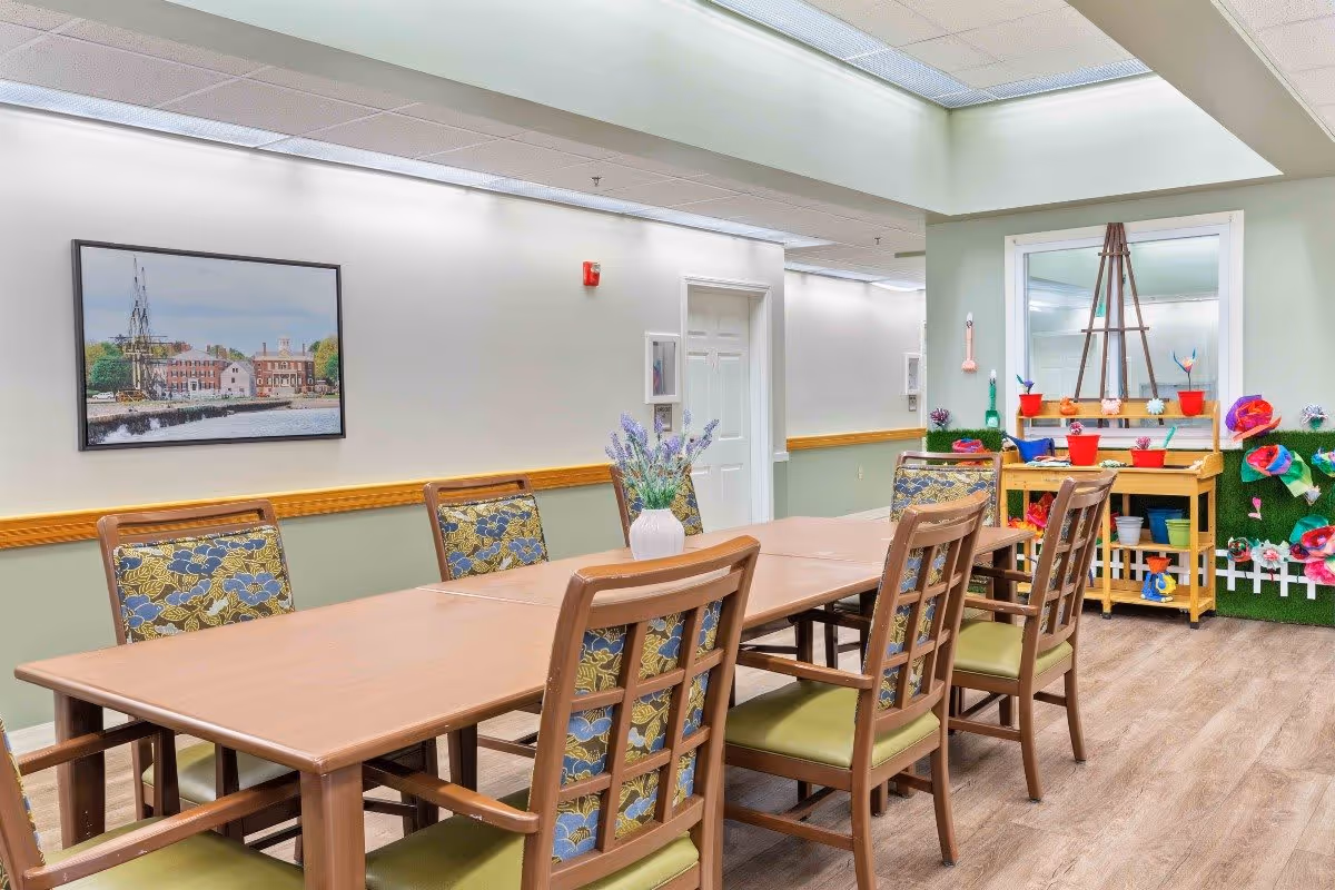 A well-lit room with a long wooden table surrounded by eight chairs with floral patterned cushions. On the table is a white vase with purple flowers. The walls are painted light green and white, with a framed picture of a waterfront scene hanging on one wall. In the background, there is a small wooden cart with colorful flower pots and decorations, and a large mirror above it. The floor is wood-patterned, and the ceiling has recessed lighting.