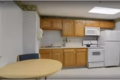 A small kitchen area with wooden cabinets, a white microwave, stove, and refrigerator. There is a round table with two chairs in the foreground. The walls have a light patterned wallpaper with a decorative border near the ceiling.