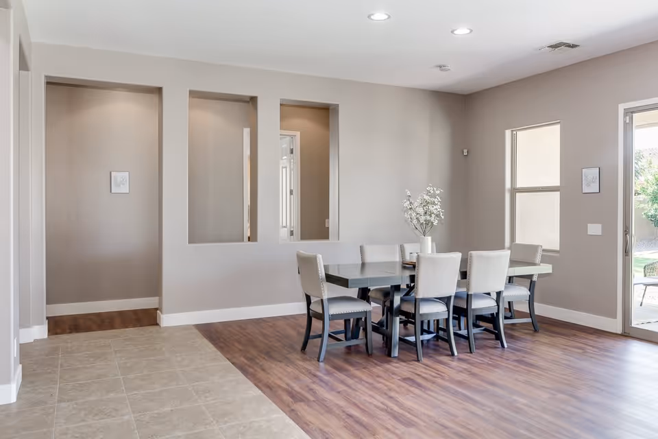 A bright dining area with a rectangular table surrounded by six upholstered chairs. The floor is a combination of light-colored tile and wood. There are three rectangular cutouts in the wall, a window, and a glass door leading outside. A vase with white flowers is placed on the table.