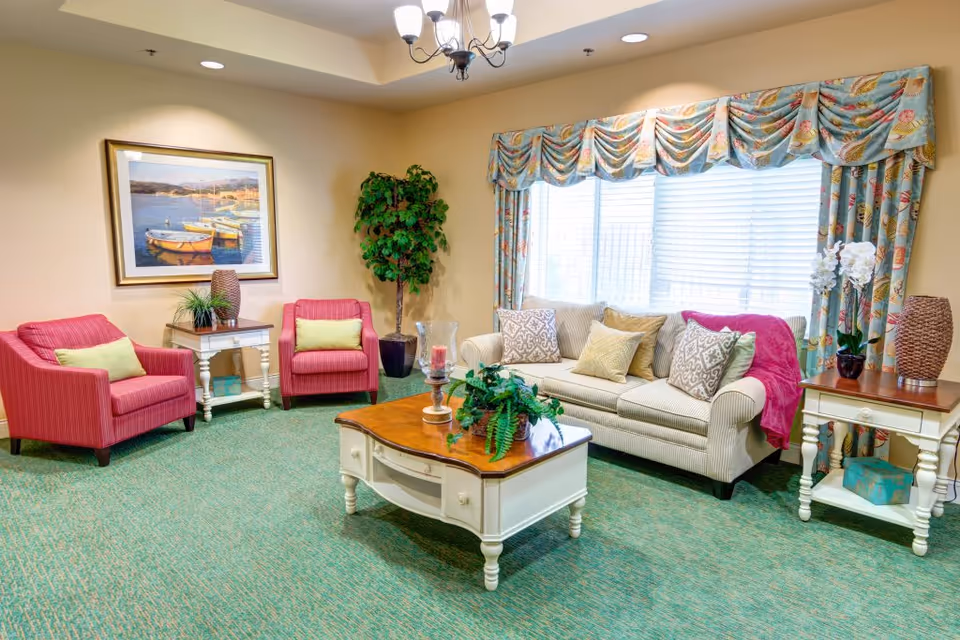 Brightly lit communal sitting area with a sofa, two pink armchairs, a coffee table, side tables, and a large window with patterned curtains.