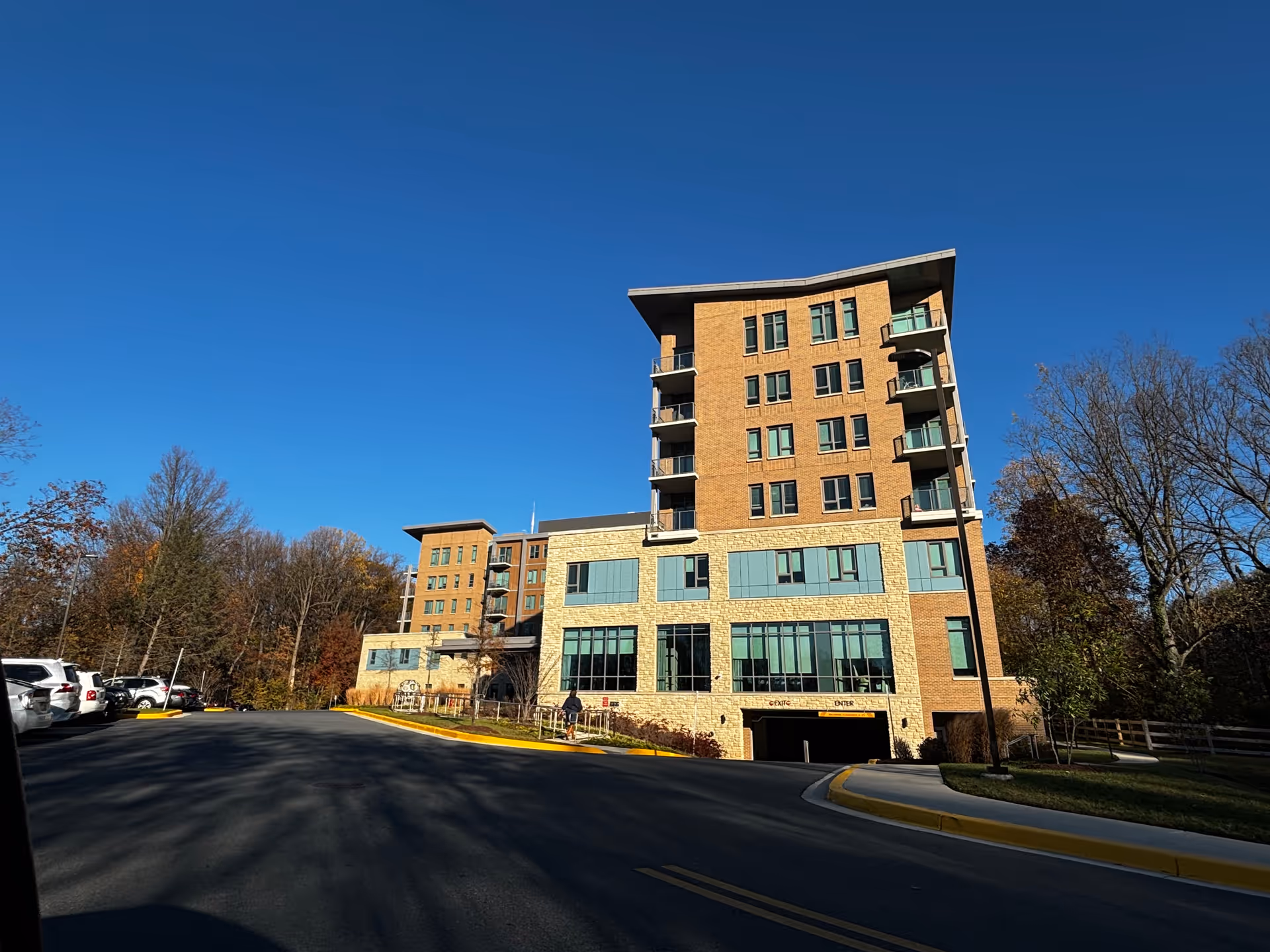 Exterior view of a multi-story senior living facility building named The Seneca Rockville, with a clear blue sky and surrounding trees. There is a driveway leading to an underground parking entrance with exit and enter signs, and several parked cars on the left side.