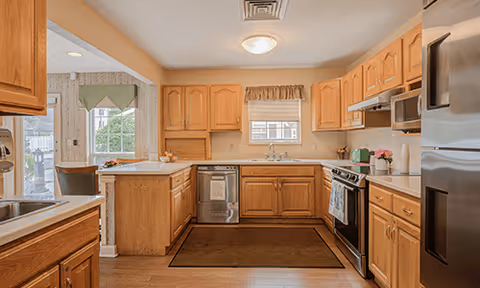 A spacious kitchen with wooden cabinets, a stainless steel refrigerator, stove, microwave, and dishwasher. There is a window above the sink with a valance, a brown rug on the floor, and an adjacent dining area visible through an open doorway.