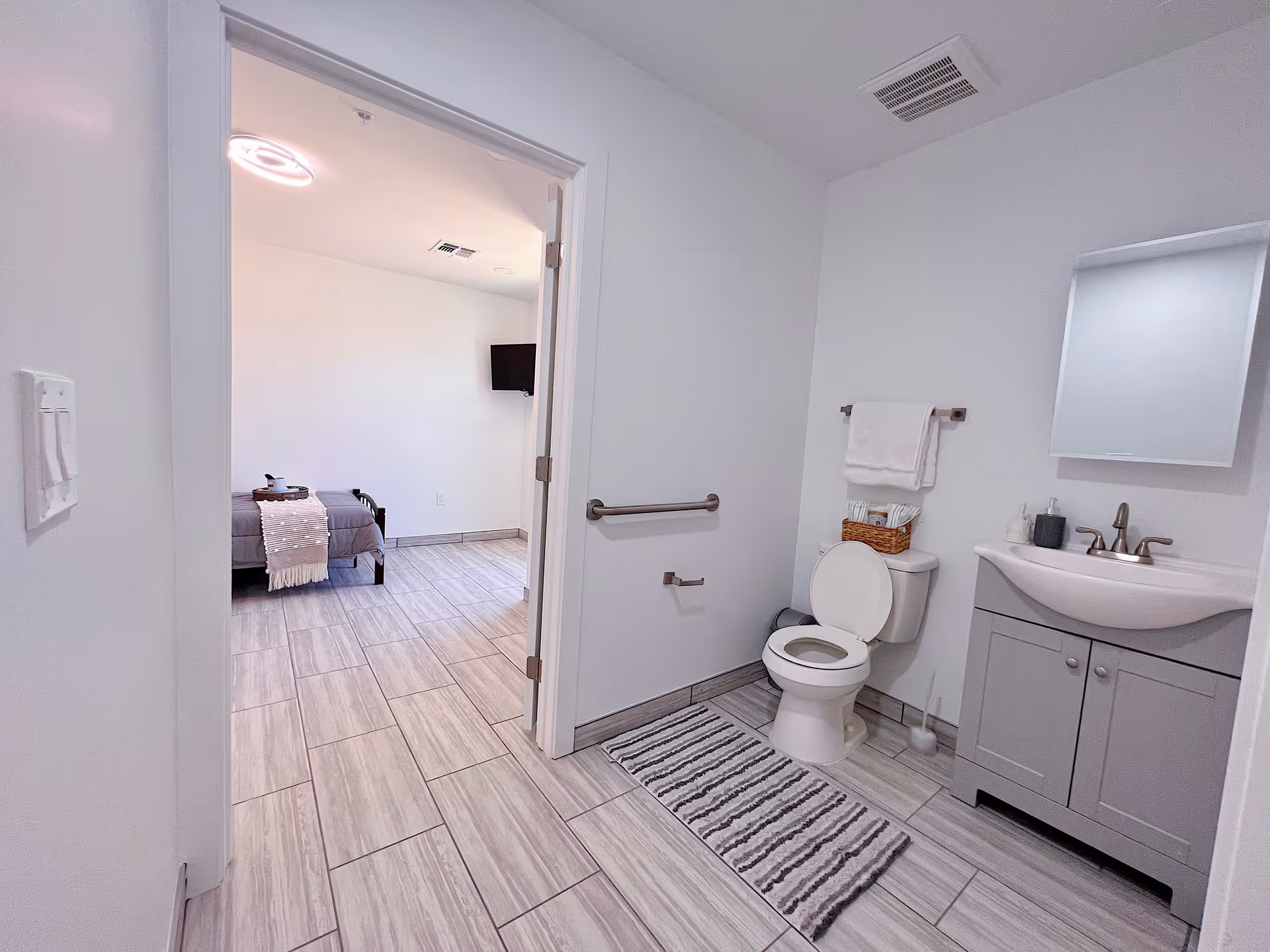 View of a bathroom with a toilet, sink with a mirror above it, and a towel rack with towels. The bathroom has light gray tiled flooring and white walls. Through an open door, a bedroom with a bed, a blanket, and a wall-mounted TV is visible.