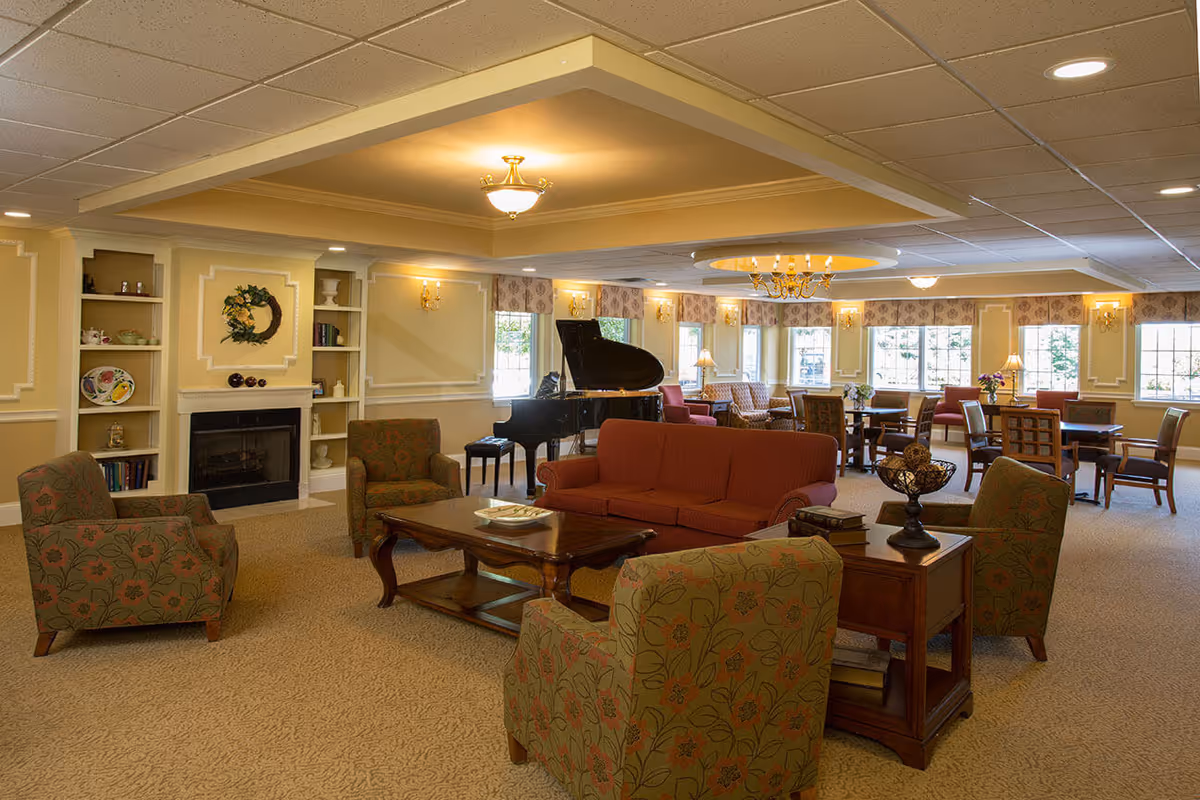 A spacious and well-lit common living area with beige walls and carpet. The room features a red sofa, several patterned armchairs, a wooden coffee table, and side tables. There is a black grand piano near the windows, built-in shelves with decorative items, and a fireplace. Multiple windows allow natural light to fill the room, and a chandelier hangs from the ceiling.