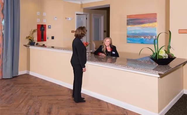 A woman standing at a reception desk talking to a receptionist seated behind the counter in a facility lobby. The reception desk has a granite countertop with a decorative plant on it. The walls are painted beige and there is a colorful abstract painting hanging on the wall behind the receptionist.