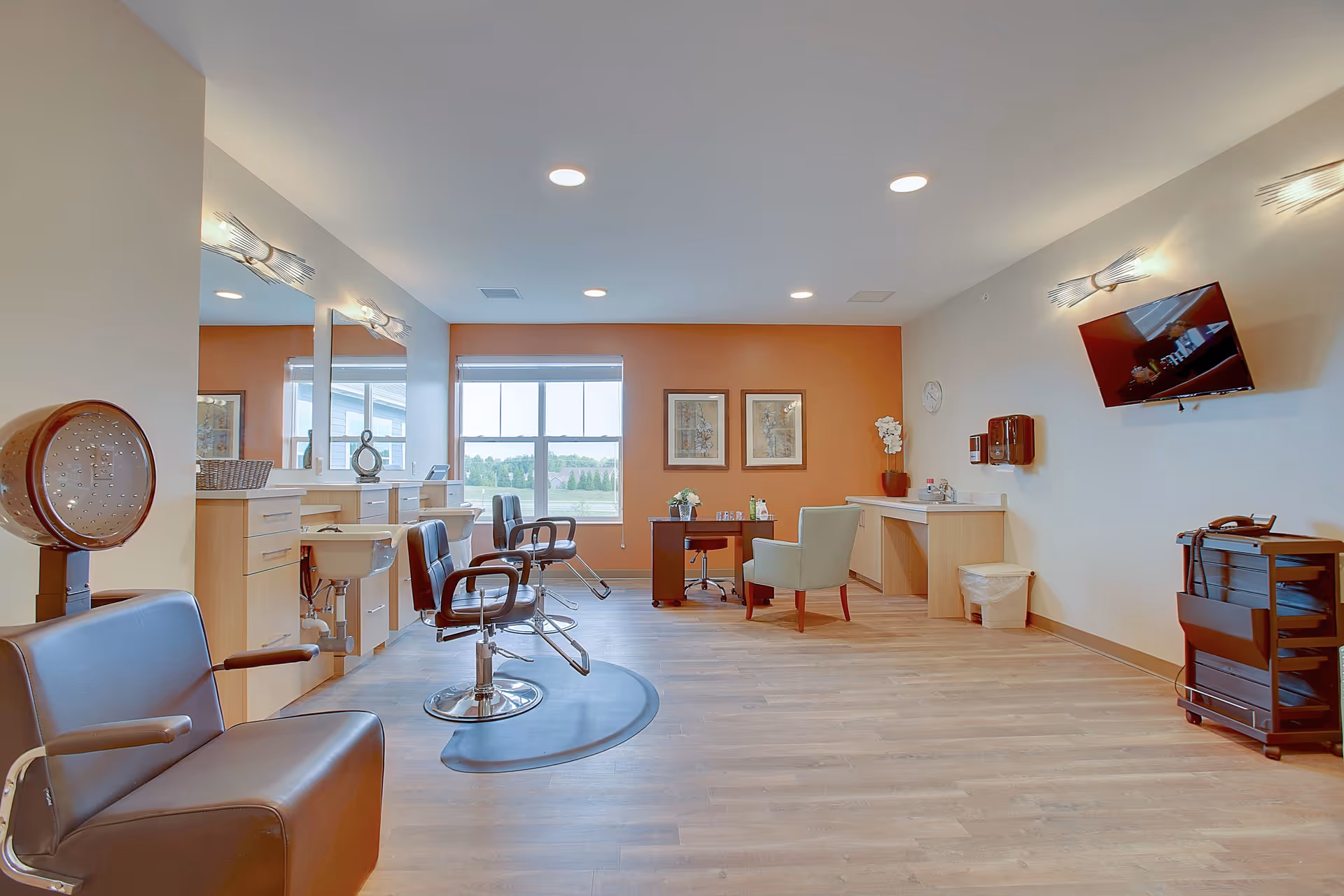 A bright and spacious salon area in a senior living facility with several black salon chairs, mirrors, and hair washing sinks along one wall. The room has light wood flooring, an orange accent wall with two framed pictures, a window letting in natural light, a small desk with a chair, a wall-mounted TV, and modern light fixtures.