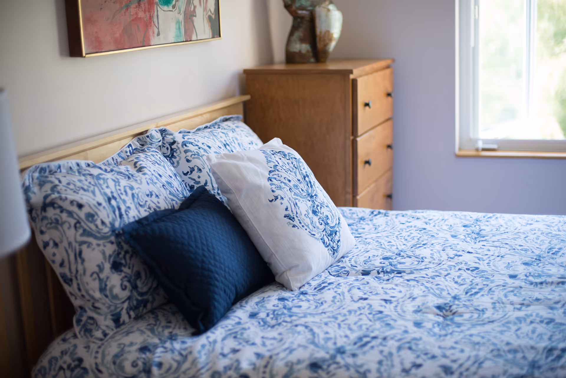 A neatly made bed with blue and white patterned bedding and pillows in a bedroom. There is a wooden dresser with decorative vases on top and a window letting in natural light.