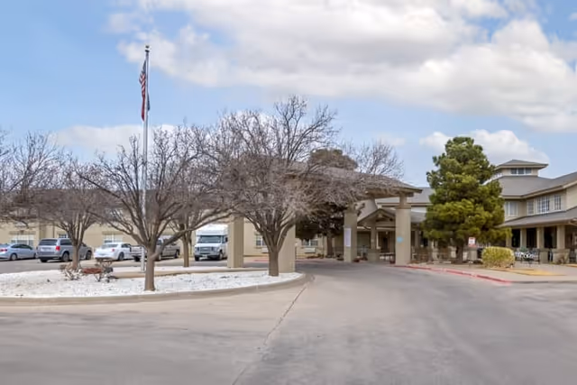 Exterior view of Brookdale Sherwood senior living facility showing the driveway entrance with a covered drop-off area, leafless trees in a circular landscaped island with white gravel, parked cars, and a flagpole with an American flag.