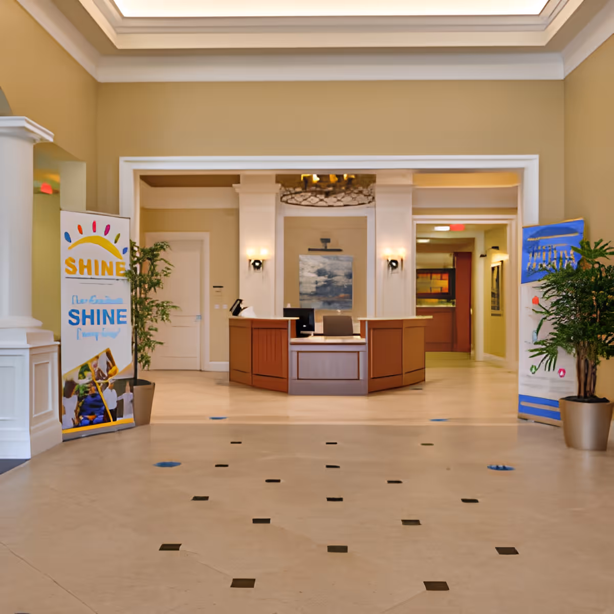 Reception area of a senior living facility with a wooden front desk, beige walls, decorative plants on either side, and banners with colorful designs and the word 'SHINE'. There is a painting on the wall behind the desk and soft lighting from wall sconces.