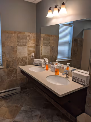Double-sink bathroom vanity with two mirrors, tiled walls and floor, soap dispensers, tissue boxes, and a window with blinds.