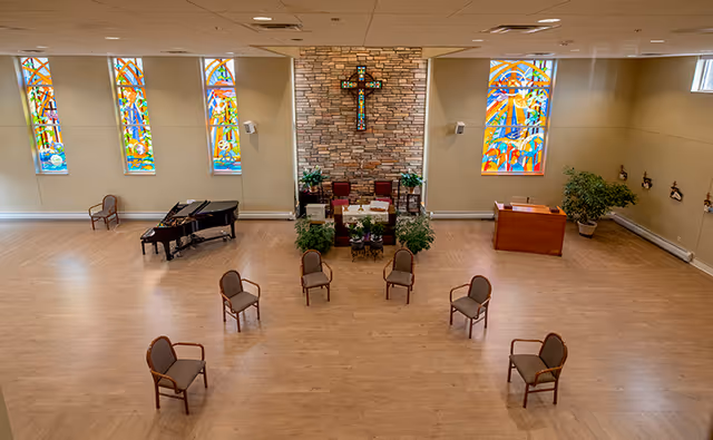 Spacious chapel-style room with a central altar and cross on a stone wall, stained glass windows, a piano, and chairs spaced across a wood floor.