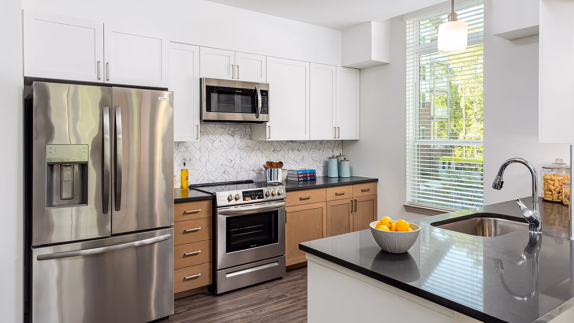 Modern kitchen with stainless steel refrigerator, oven, and microwave. The kitchen features white upper cabinets, light brown lower cabinets, a black countertop, a sink with a chrome faucet, a bowl of lemons on the counter, and a large window with white blinds letting in natural light.