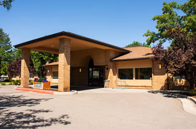 Exterior view of a single-story brick building with a covered entrance supported by four brick columns. There are trees and plants around the building, and a clear blue sky above.