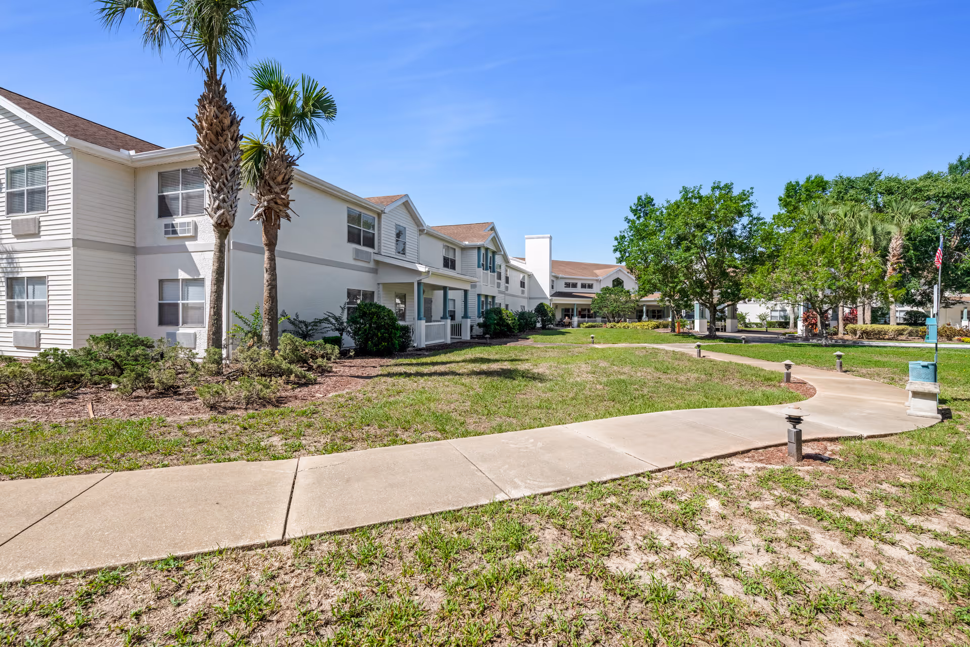 Exterior view of a senior living facility with a two-story white building, palm trees, green grass, and a curved concrete walkway leading towards the entrance under a clear blue sky.