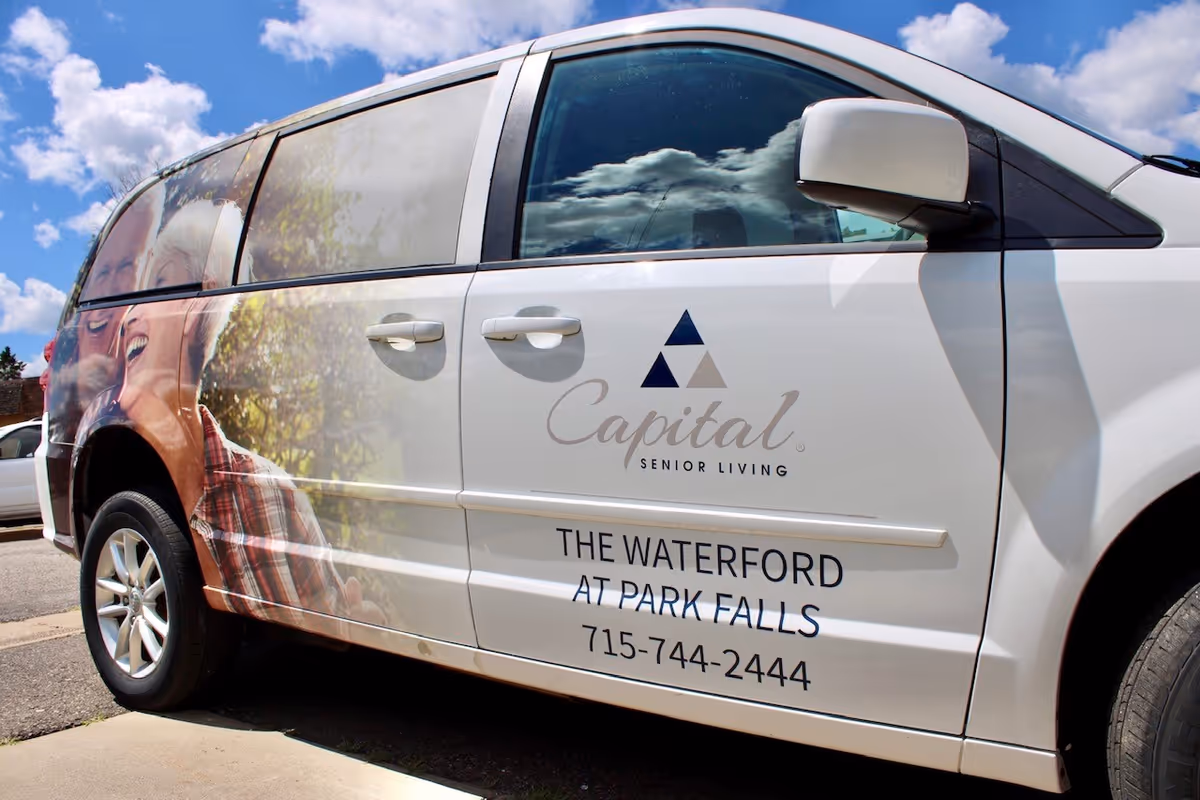 Branded white minivan for The Waterford at Park Falls with Capital Senior Living logo parked under a blue sky.