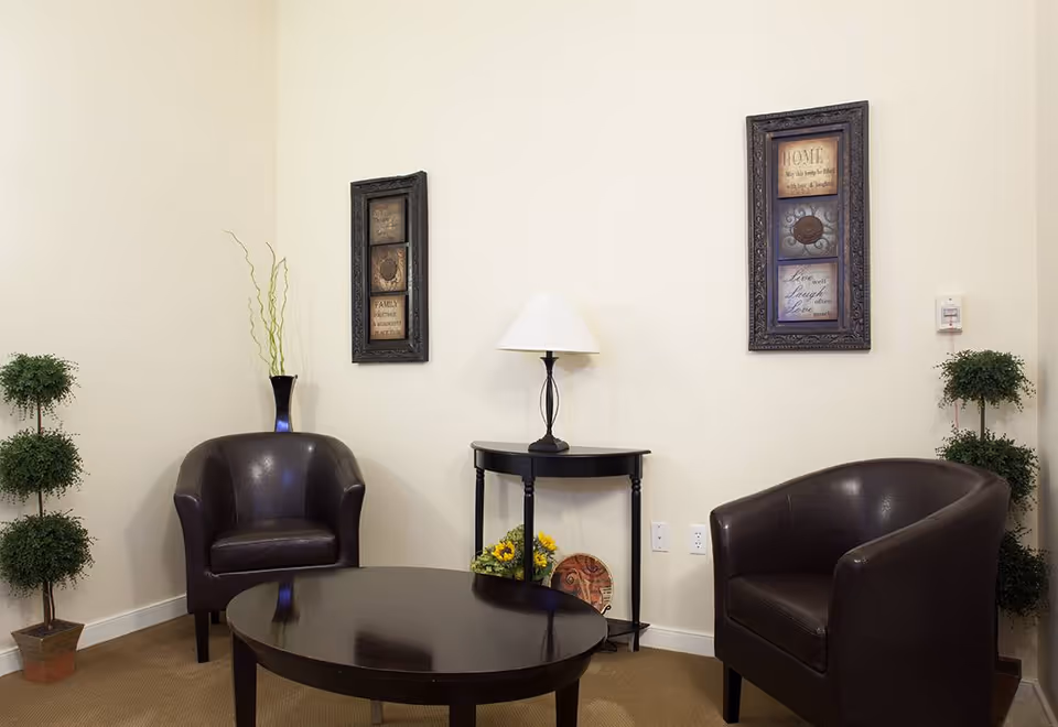 A cozy sitting area with two dark brown leather armchairs facing a round dark wooden coffee table. Between the chairs is a small black side table with a white lampshade and a small flower arrangement underneath. Two framed decorative wall hangings are mounted on the light beige wall behind the chairs. Two potted topiary plants are placed in the corners of the room.