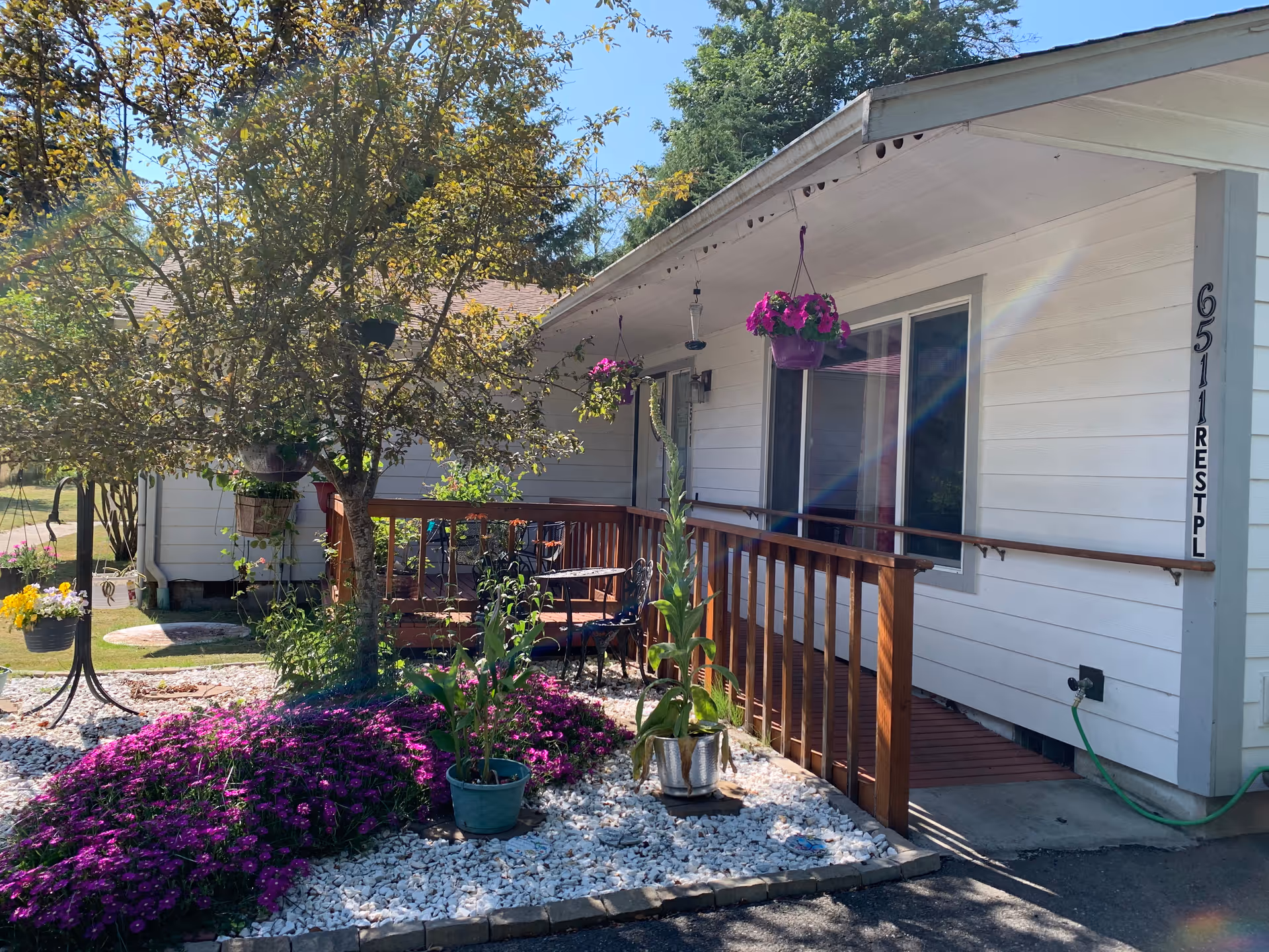 Exterior view of a white single-story building with a wooden ramp and railing leading to the entrance. There are hanging flower pots with pink flowers and a garden area with purple flowers and green plants surrounded by white gravel. A tree is also visible in the garden area under a clear blue sky.