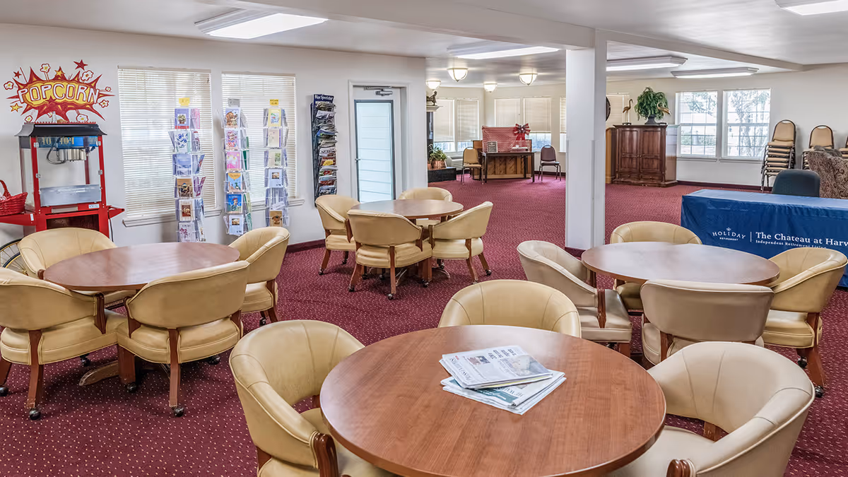 Interior view of a common area with round wooden tables surrounded by beige cushioned chairs on a red carpet. There is a popcorn machine on the left side near windows with blinds, and racks holding greeting cards. In the background, there is a blue tablecloth with the text 'Holiday | The Chateau at Harveston' and some stacked chairs near large windows.
