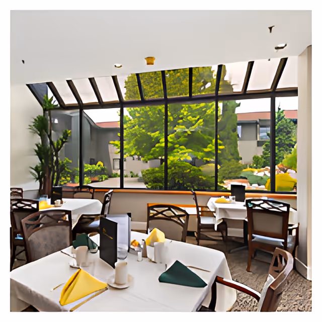 Dining area with tables covered in white tablecloths, set with folded yellow and green napkins, cups, and menus. Large windows and skylights provide a view of green trees and outdoor landscaping.