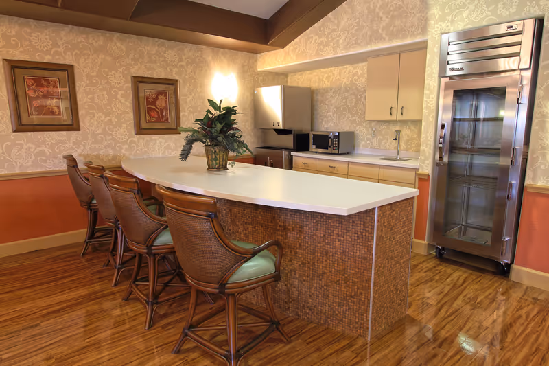 A kitchen area with a long white countertop island surrounded by four wooden chairs with green cushions. The back wall has beige cabinets, a microwave, a small sink, and a large stainless steel refrigerator. The floor is wooden, and the walls have a patterned wallpaper with two framed pictures and a wall-mounted light fixture. A potted plant is placed on the countertop.