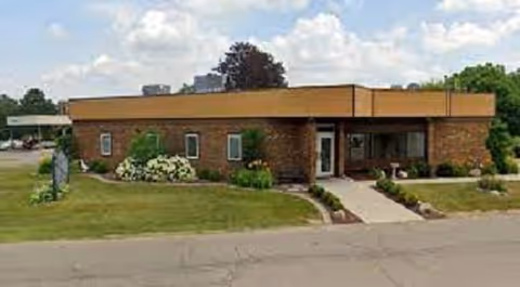 Single-story brick building with a flat roof, surrounded by grass and some shrubs, under a partly cloudy sky.
