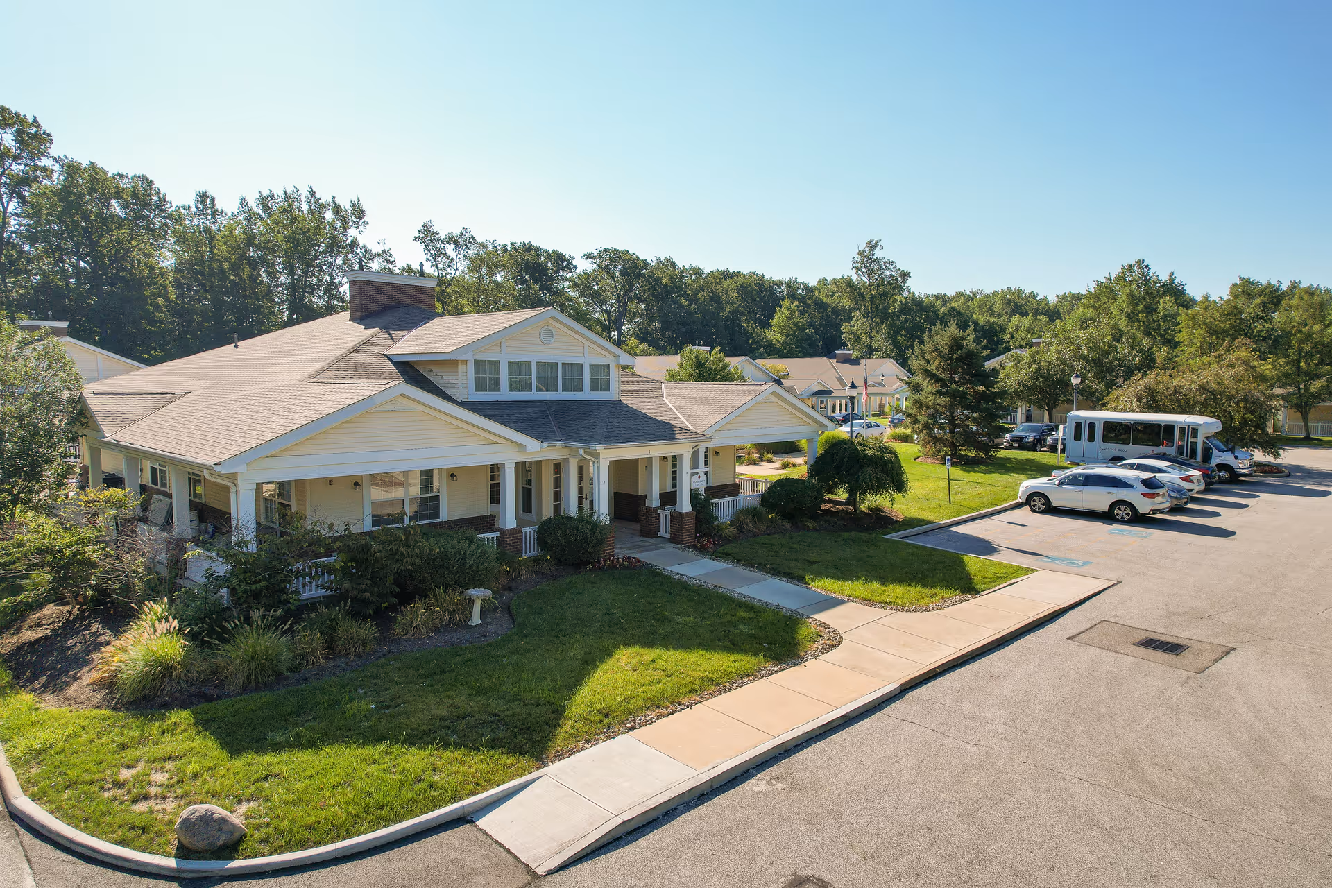 Exterior view of a single-story senior living facility building with a beige roof and white siding, surrounded by green trees and landscaped bushes. There is a parking lot with several cars and a shuttle bus parked nearby under a clear blue sky.