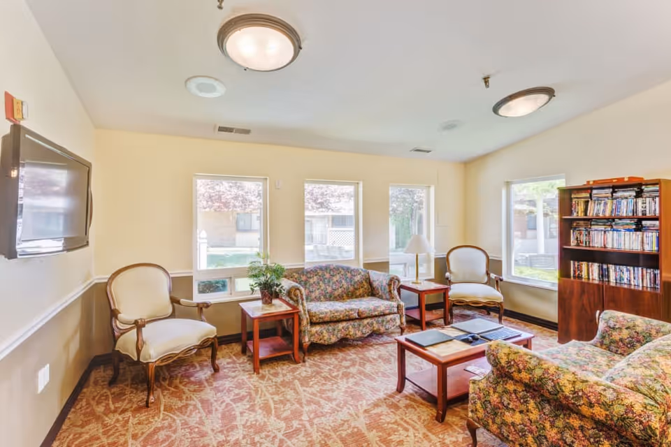 A cozy living room with floral patterned sofas, two armchairs, a wooden coffee table with books and a remote, two side tables with a lamp and a plant, a bookshelf filled with DVDs, and three windows letting in natural light.