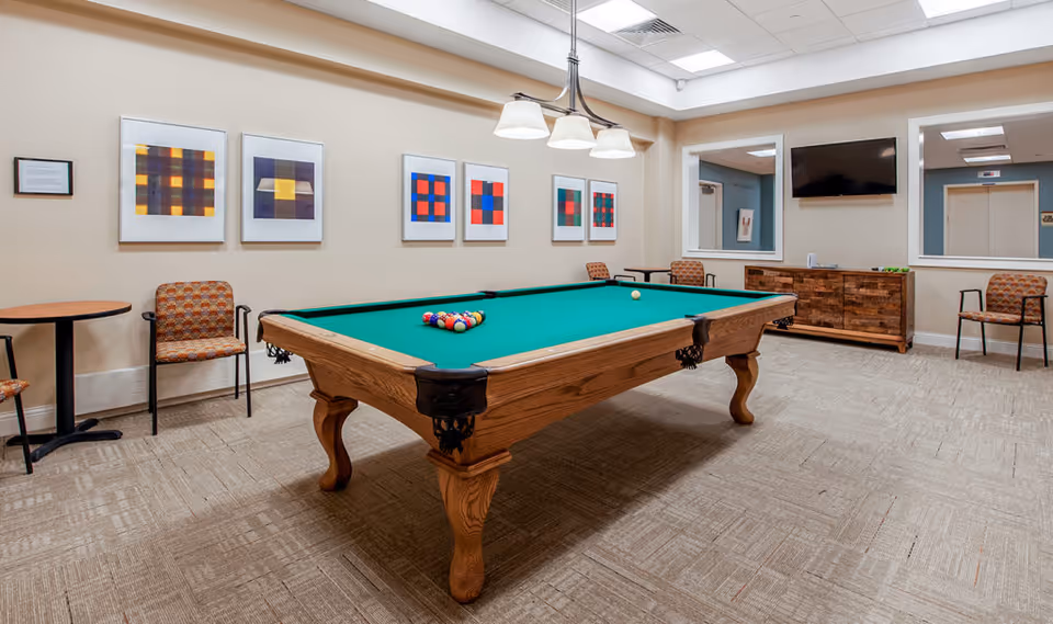 A well-lit game room featuring a wooden pool table with a green felt surface and a full set of billiard balls arranged for a game. The room has beige walls adorned with colorful abstract artwork in white frames. There are several patterned chairs and small round tables along the walls. A wooden cabinet with a flat-screen TV mounted above it is visible, along with two large mirrors reflecting parts of the room.