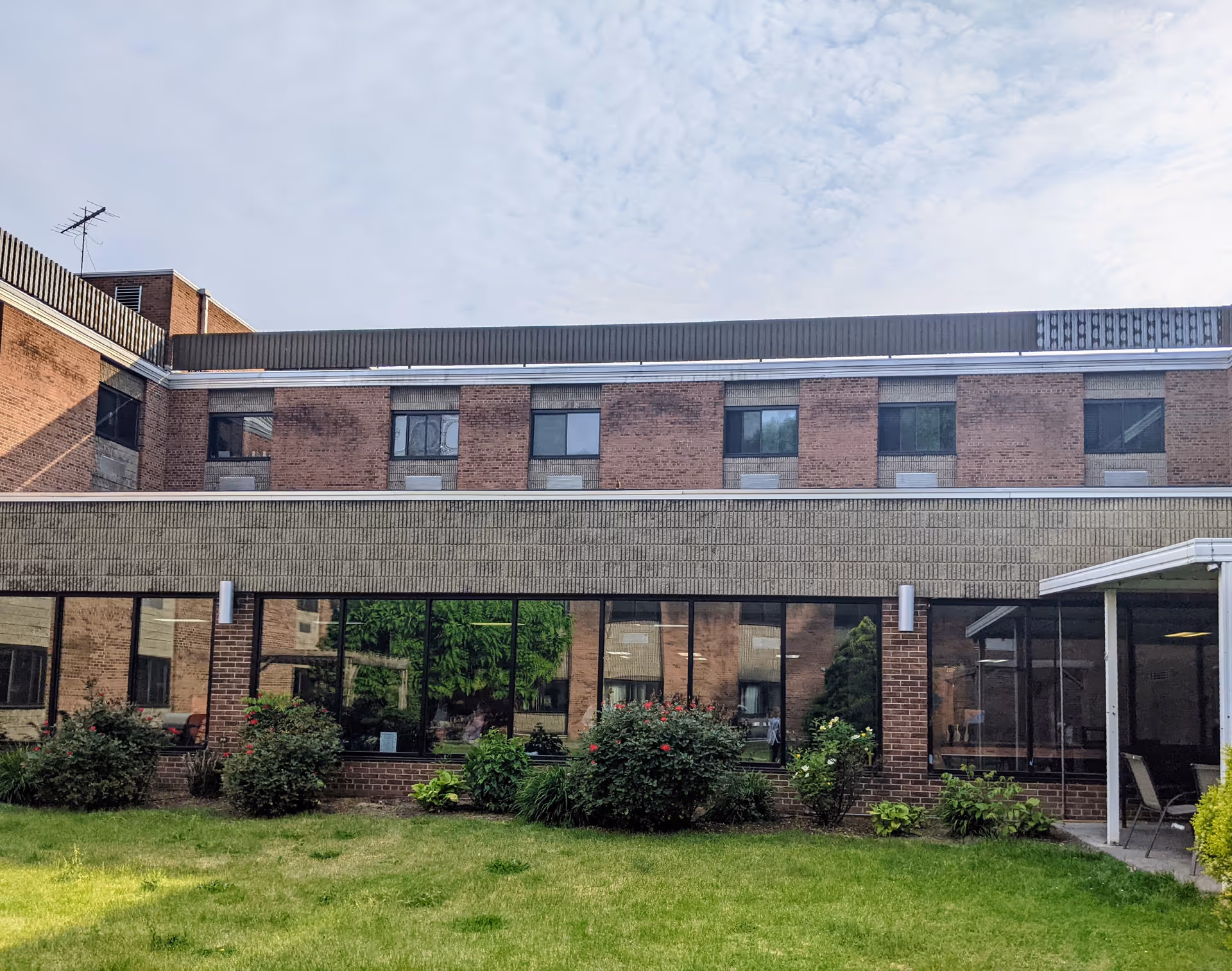 Exterior view of a multi-story brick building with large windows on the ground floor and smaller windows on the upper floors. There is a grassy lawn with bushes and plants in front of the building under a partly cloudy sky.