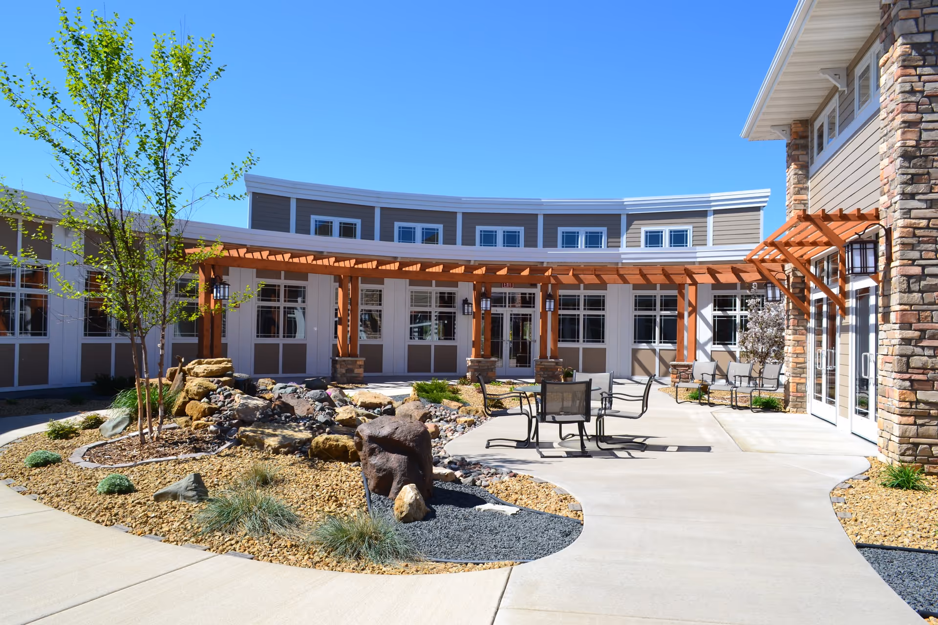 Outdoor courtyard area of a senior living facility with a curved building facade, a wooden pergola, patio tables and chairs, landscaped garden with rocks and small plants, and a clear blue sky.