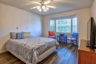 A bedroom with a bed covered in a blue and white patterned bedspread, two colorful chairs and a small table near a large window, a ceiling fan with lights, and a flat-screen TV on a wooden dresser.