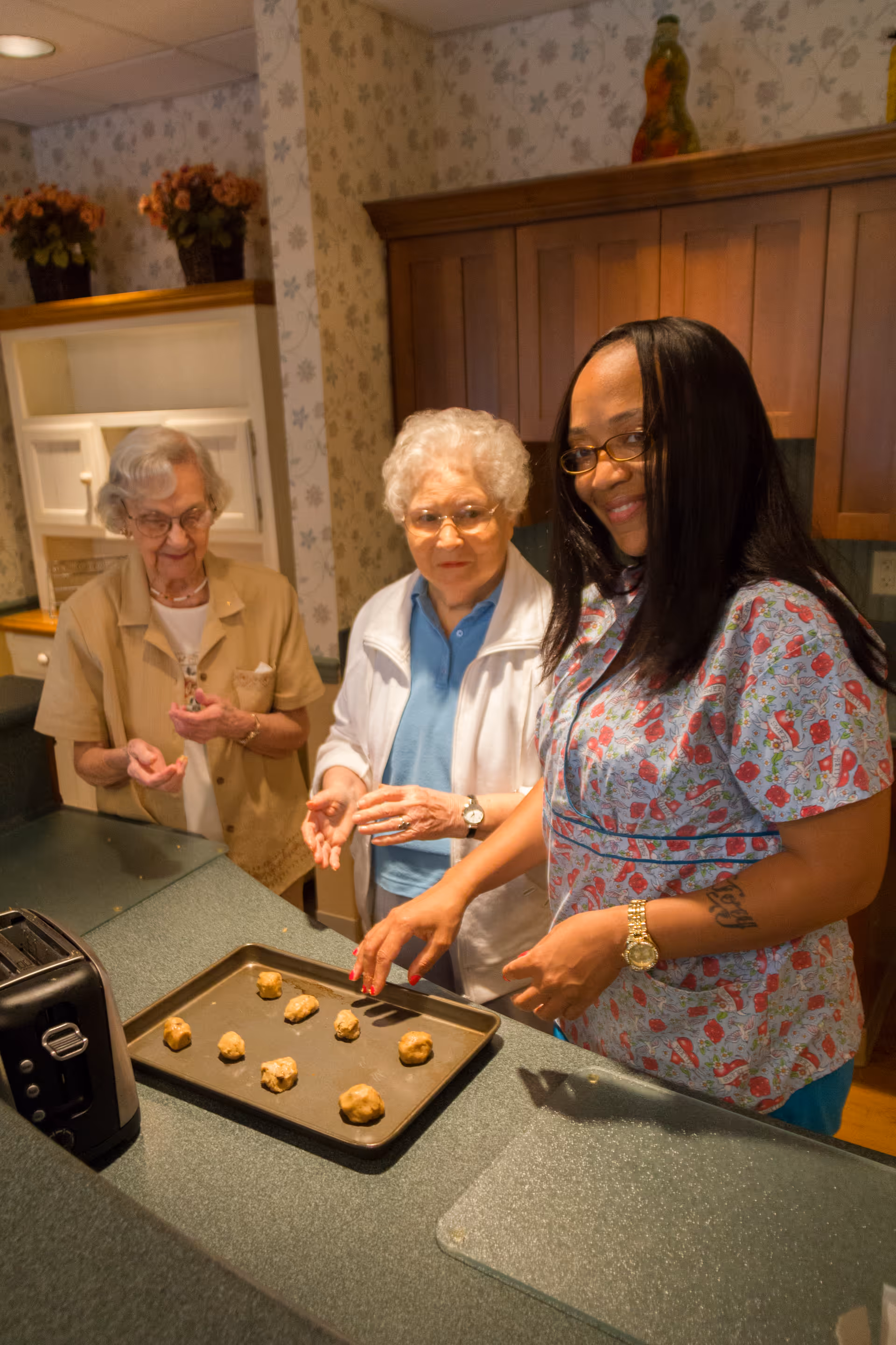 Three women in a kitchen preparing cookie dough on a baking tray. Two elderly women and one younger woman are smiling and placing dough balls on the tray. The kitchen has wooden cabinets and floral wallpaper.