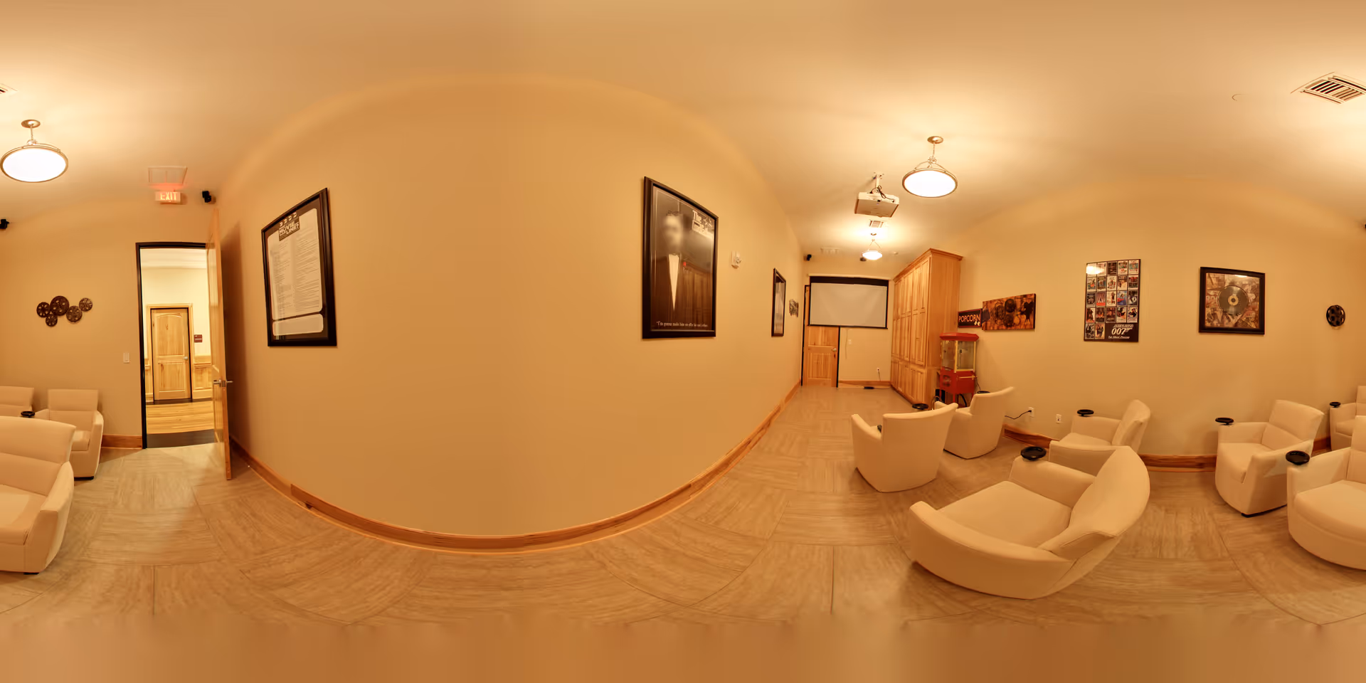 A cozy entertainment room with several beige armchairs arranged facing a projector screen on the far wall. The room has warm lighting, light-colored walls, and wood trim. Movie posters and framed pictures decorate the walls. A popcorn machine is visible near the screen, and the floor is covered with light-colored tiles.